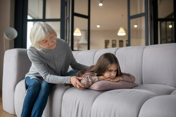 Little offended girl lying on the couch and grandmother sitting nearby.