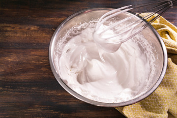 Process of cooking meringue with whisk in metal bowl on wooden table. Recipe with whipped eggs, sugar. Perfect peaks for white sweet cookies, Italian or French dessert. Flat lay, copy space.