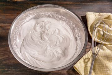 Process of cooking meringue with whisk in metal bowl on wooden table. Recipe with whipped eggs, sugar. Perfect peaks for white sweet cookies, Italian or French dessert. Flat lay, copy space.