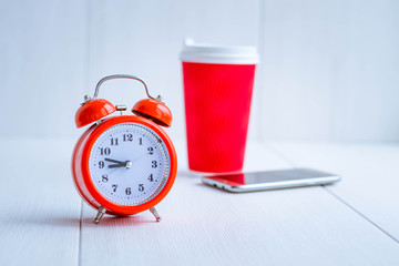 Orange alarm clock in the foreground. Red paper coffee cup with white plastic lid and mobile phone....