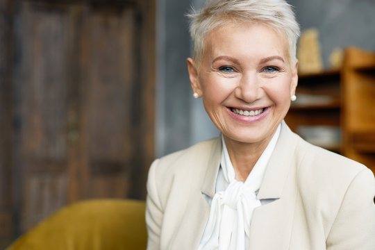 Mature People, Aging, Business And Occupation Concept. Beautiful Sixty Year Old Female Wearing Pearl Earrings, Beige Jacket Over White Blouse Working In Her Office, Having Joyful Positive Look