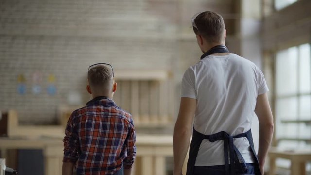 Back view follow shot of carpenter and his little son walking towards workbench in carpentry shop in slow motion and discussing construction of large wooden boat model
