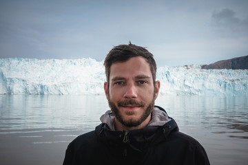Happy and smiling young man standing in front of Eqip Sermia glacier called Eqi Glacier. Tourist with huge wall of ice in background. The concept of global warming and professional guides. Overcast