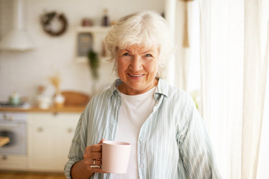 Positive friendy looking senior elderly woman with gray hair and wrinkles spending day at home, posing by window with cozy clean kitchen in background, drinking tea or coffee in the morning