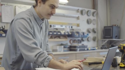 Medium arc shot of Caucasian male artisan with long hair in bun planning new furniture design, using laptop and preparing technical drawings while working at carpentry workshop