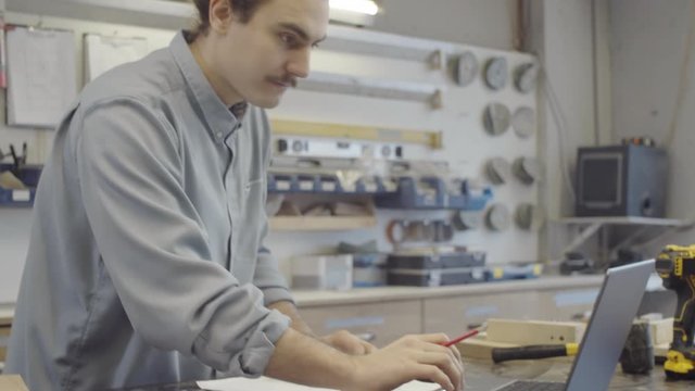 Medium arc shot of Caucasian male artisan with long hair in bun planning new furniture design, using laptop and preparing technical drawings while working at carpentry workshop