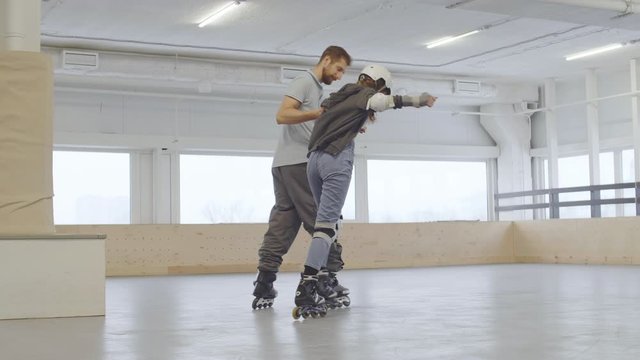Wide shot of male coach holding female novice rollerblader wearing protecting gear and trying to skate