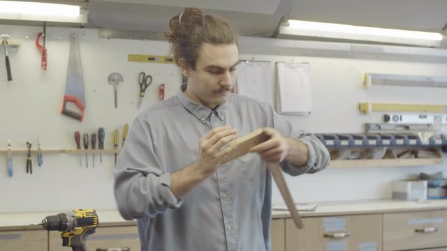Waist-up shot of young Caucasian male carpenter with long hair in bun and moustache working in workshop and joining together wooden furniture components with mortise and tenon joint