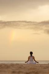 Yoga lotus pose meditation practice of young female on a beach with rainbow