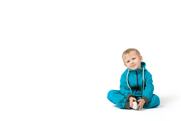 The baby blond-haired boy sits on the floor in a turquoise jumpsuit and smiles at the camera on a white background with place for text.
