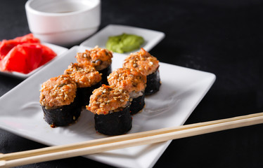 6 baked rolls on a terelka, saucers with pickled ginger and wasabi, sticks on a black background, Japanese rolls with fish and sesame seeds