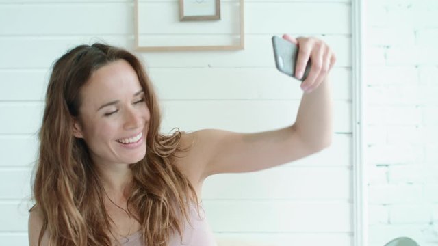 Portrait of smiling pregnant woman taking selfie photo in light bedroom.