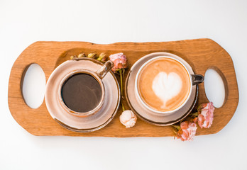Two cups of coffee decorated with hearts on a wooden tray decorated with roses are on a white table