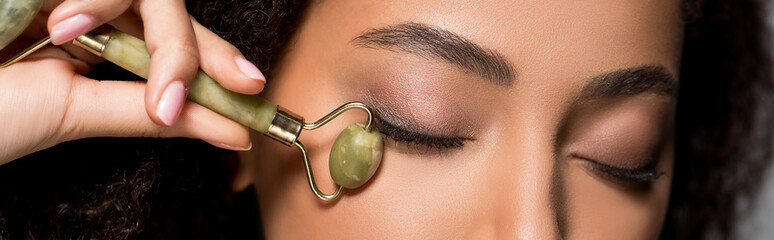 panoramic shot of african american girl with closed eyes using jade roller, on grey