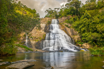 Young woman in a Sunny day in the Tropical waterfall falls from the mountain cliff to the jungle, serene landscape of Diyaluma falls.
