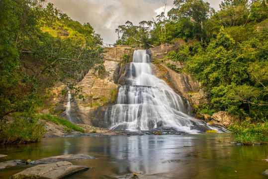 Young woman in a Sunny day in the Tropical waterfall falls from the mountain cliff to the jungle, serene landscape of Diyaluma falls.