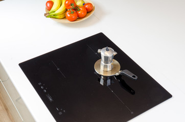  induction cooker with an adapter, coffee tea pot mokka coffee maker, modern glass kitchen stove top, low energy consumption and high efficiency, black glass on white kitchen worktop, fruit on te plat