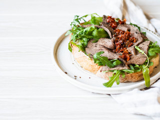 Large toast on homemade bread with soft cheese, arugula and slices of roast beef with sun-dried tomatoes on top. Copy space.