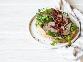 Large toast on homemade bread with soft cheese, arugula and slices of roast beef with sun-dried tomatoes on top. Copy space.