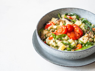 Salad of quinoa, baked vegetables, spinach and raw cucumber in a blue plate on a white wood background