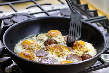 Fried eggs with sausage and seasonings in a black pan on a gas stove. Tasty and nutritious breakfast for the family. Selective focus. Closeup view