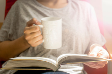 Woman with cup of hot beverage reading book at home in winter evening, closeup