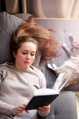 Attractive female with long hair reading interesting book, resting with cat at home