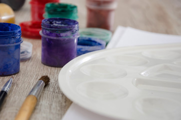 colored gouache in jars, a brush and a palette for drawing on a wooden table.