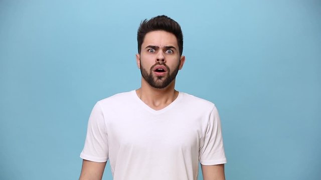 Handsome unshaven bearded young guy 20s wearing white t-shirt isolated over pastel blue background in studio. People sincere emotions lifestyle concept. Looking at camera scared shocked frightened