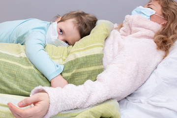 Girl and child are lying on the bed in medical masks. Virus, mom is sick and daughter hugs mom. European Family and Coronavirus