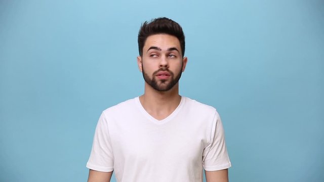 Handsome unshaven bearded young guy 20s wearing white t-shirt isolated on pastel blue background in studio. People sincere emotions lifestyle concept. Looking in different sides directions suspicious
