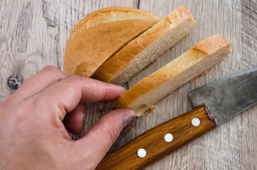 hand takes a slice of loaf. White bread and a knife on a wooden table. View from above.