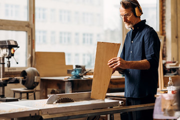 Male carpenter wearing noise-canceling headphones and holding a wood plank