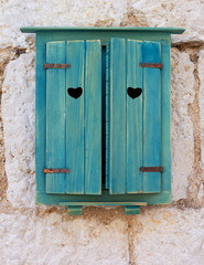 The window is covered with blue wooden shutters made of boards with decorative holes in the form of hearts in an old stone house