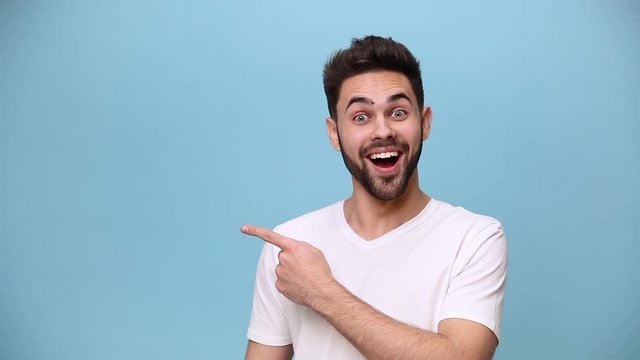 Handsome charismatic unshaven bearded young guy 20s wearing white t-shirt isolated over pastel blue background in studio. People sincere emotions lifestyle concept Pointing fingers on copy space aside