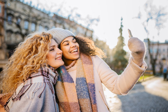 always online concept. Emotional portrait of excited african female talking with girl friend showing smart phone outdoors in city street. Curly-hair biracial friends in scarf and jacket in winter day.