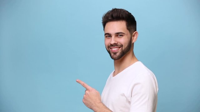 Handsome charismatic unshaven bearded young guy 20s wearing white t-shirt isolated over pastel blue background in studio. People sincere emotions lifestyle concept Pointing fingers on copy space aside