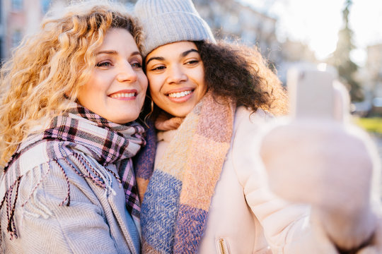 always online concept. Portrait of excited african female talking selfie portrait with girl friend outdoors in city street. Curly-hair biracial friends in scarf and jacket in sunny winter day.