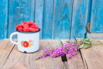Old enamel cup with handful of fresh rasperries and violet field flower on rustic wooden table. 