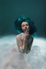 A beautiful girl with blue and long hair swims underwater in the pool in a fluffy white dress. Looks like a nymph or a mermaid
