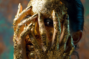 Girl with a mask on her face made of gold leaf. Gloomy studio portrait of a brunette on an abstract background.