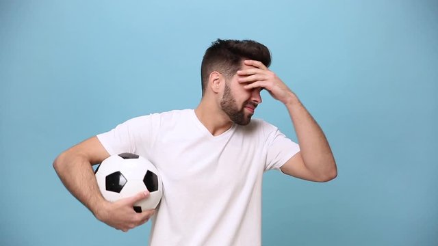 Bearded young guy 20s football fan cheer up support favorite team with soccer ball expressive gesticulating hands, in white t-shirt isolated on pastel blue background in studio. People sport concept