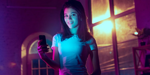 Smiling, looking forward. Cinematic portrait of stylish woman in neon lighted interior. Toned like cinema effects in purple-blue. Caucasian model using smartphone in colorful lights indoors. Flyer.