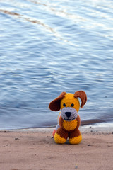 Amigurumi doll puppy on the beach in a pine forest near a forest lake