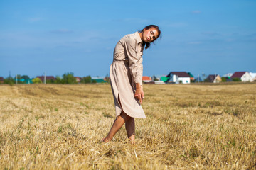 Young beautiful brunette model in a beige dress posing against the background of a mowed wheat field