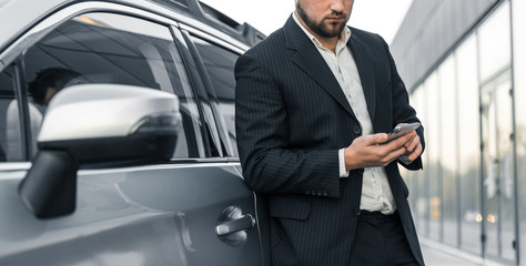 Handsome businessman with smartphone near vehicle on office parking