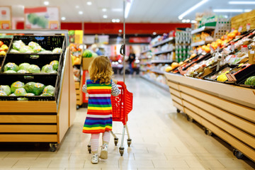 Cute todler girl pushing shopping cart in supermarket. Little child buying fruits. Kid grocery...