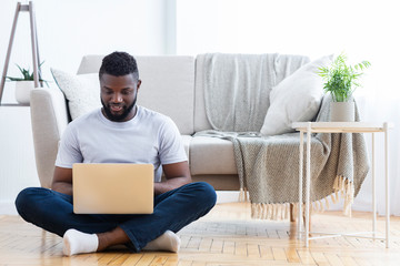 Black freelancer typing on laptop, sitting on floor at home