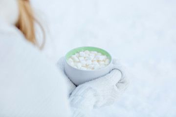 Mug of hot coffee with marshmallow in his hands in white mittens on the background of snow close up