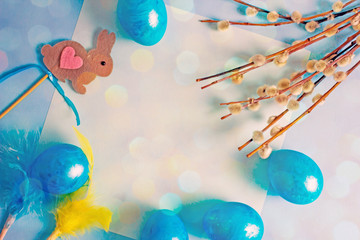 Happy Easter day. A yellow sheet of paper surrounded by blue Easter eggs, pussy-willow twigs, decorative Easter hare, flowers on a blue background, top view, copy space. Easter festive background 